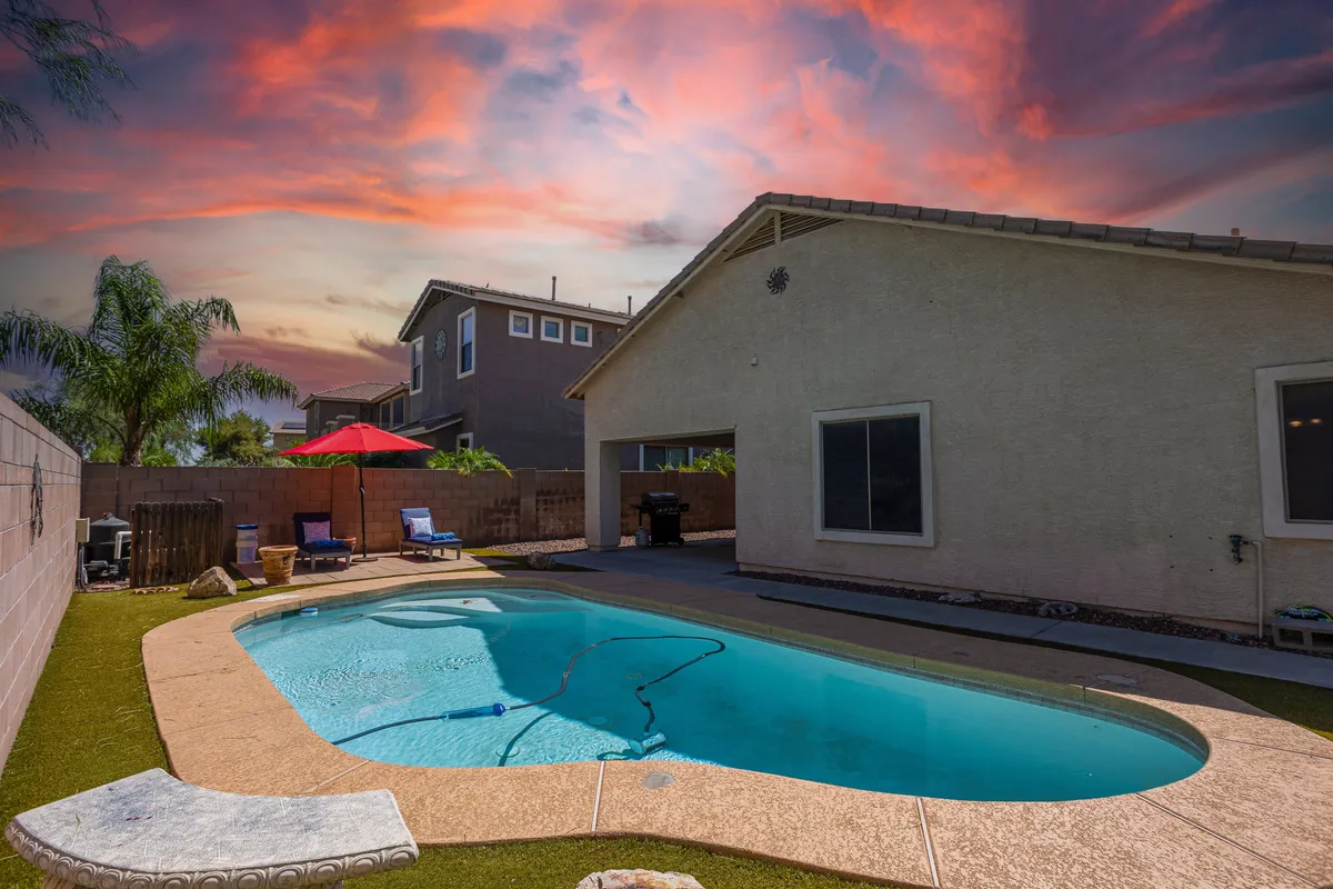 This image showcases a sweeping view of the backyard, featuring the sparkling swimming pool at the center, a welcoming patio area with seating, an umbrella for shade, and lush palm trees framing the outdoor space. The inviting atmosphere hints at both relaxation and lively social gatherings, with ample room for lounging, swimming, or enjoying a grilled meal.