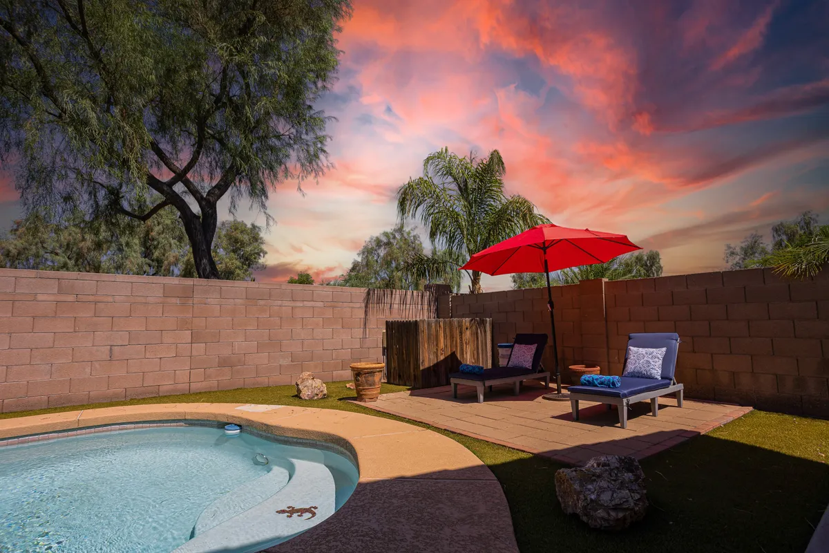 A tranquil view of the pool area with a red umbrella, several lounge chairs, lush green grass, and towering palm trees. The setting evokes peaceful moments spent reading, sunbathing, or simply soaking in the Arizona sky, with the pool at the heart of the scene.