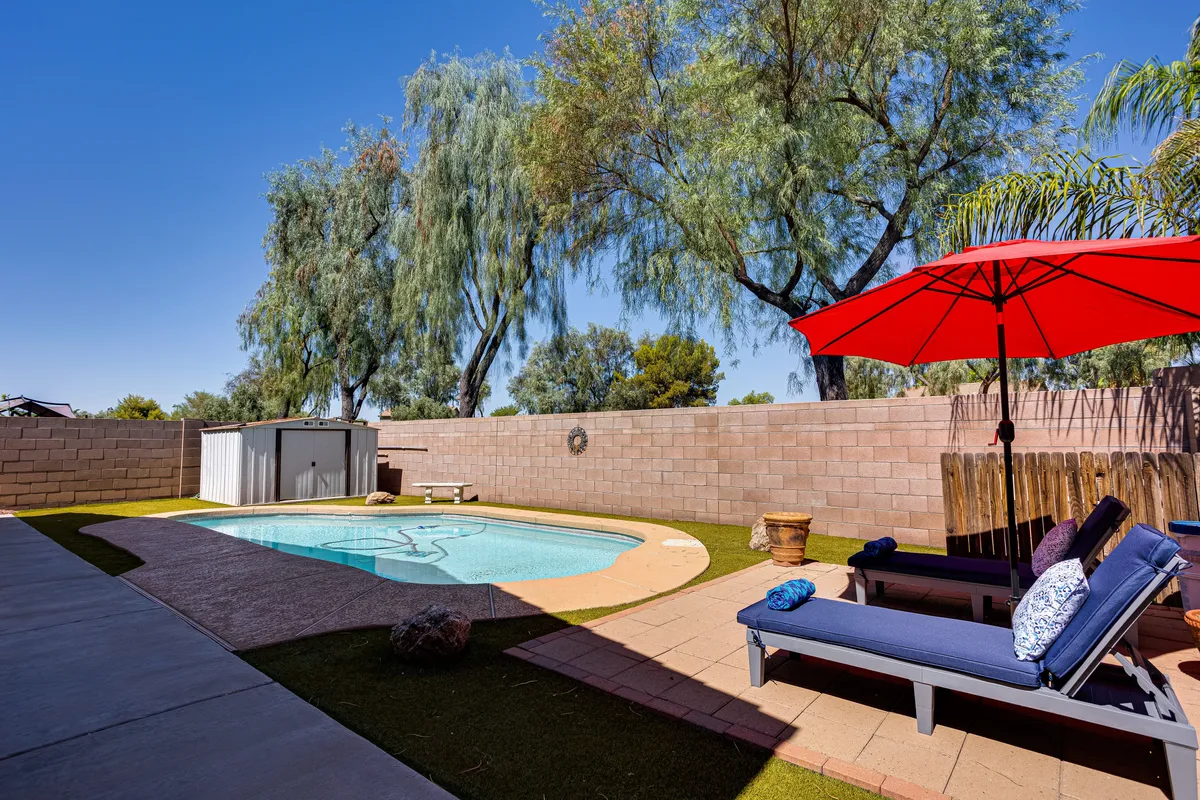 A vibrant outdoor scene highlighting the swimming pool, a prominent red umbrella, comfortable lounge chairs, and green trees. The area blends relaxation and social spaces, suggesting both serene and festive uses of the backyard.