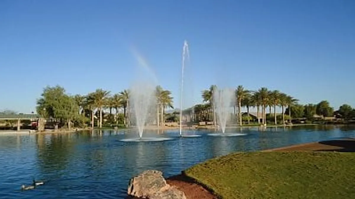 A tranquil exterior scene featuring fountains, palm trees, and calm reflective water, capturing the lush, resort-like amenities offered by the neighborhood. The setting hints at scenic vistas and a sense of retreat.