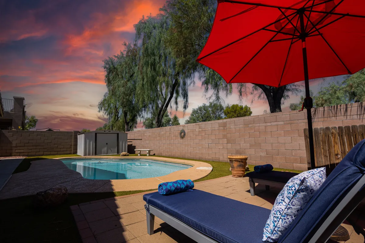 A tranquil backyard scene showcasing a sparkling swimming pool surrounded by red umbrellas and comfortable lounge chairs, inviting guests to sunbathe, swim, or simply relax. The pool area is bathed in soft sunset light, enhancing the serene and inviting mood.