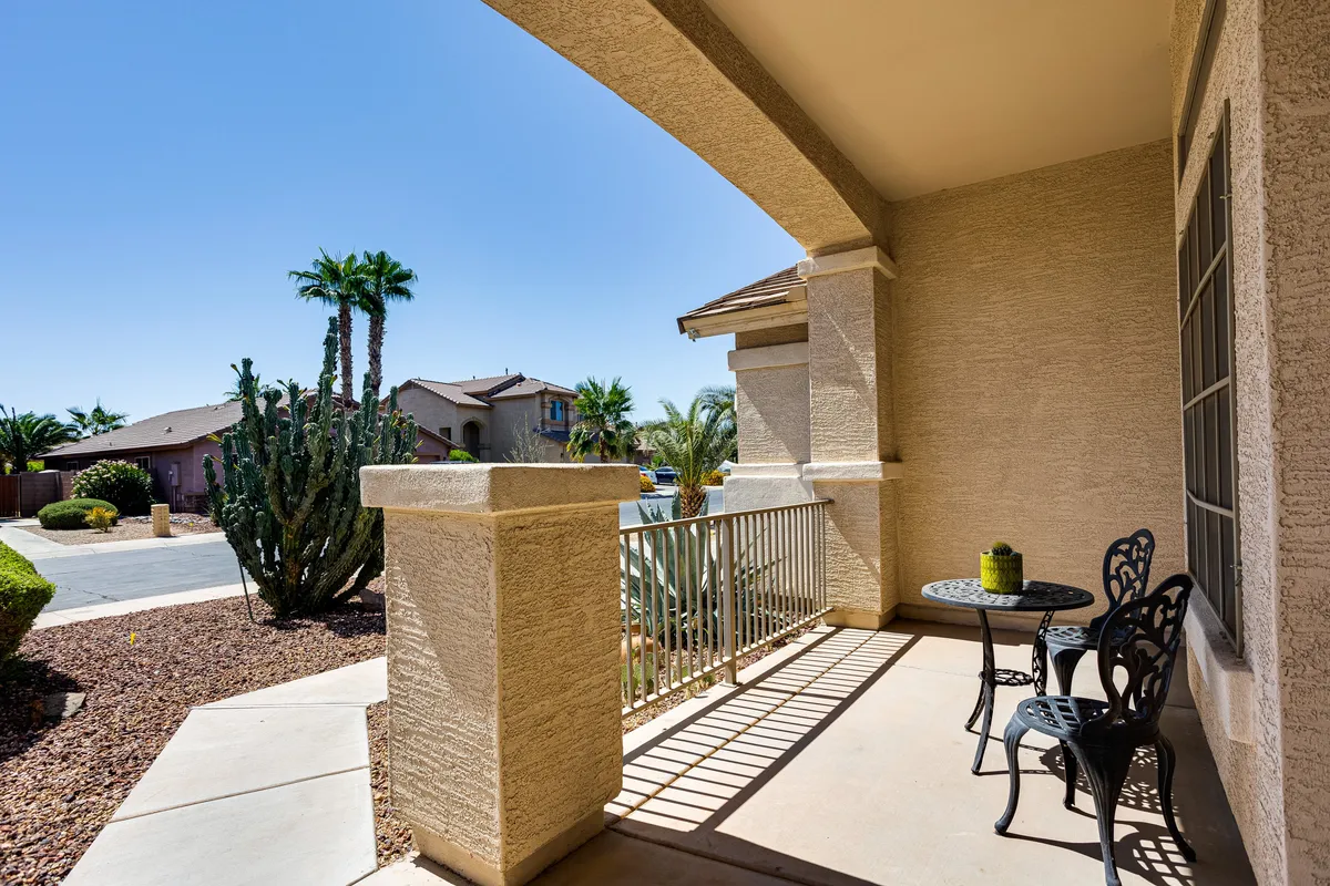 A serene shot of a small round table and two chairs on a porch, surrounded by cactus and palm trees. The outdoor scene suggests opportunities for relaxation and casual socializing, bathed in gentle morning or evening light.