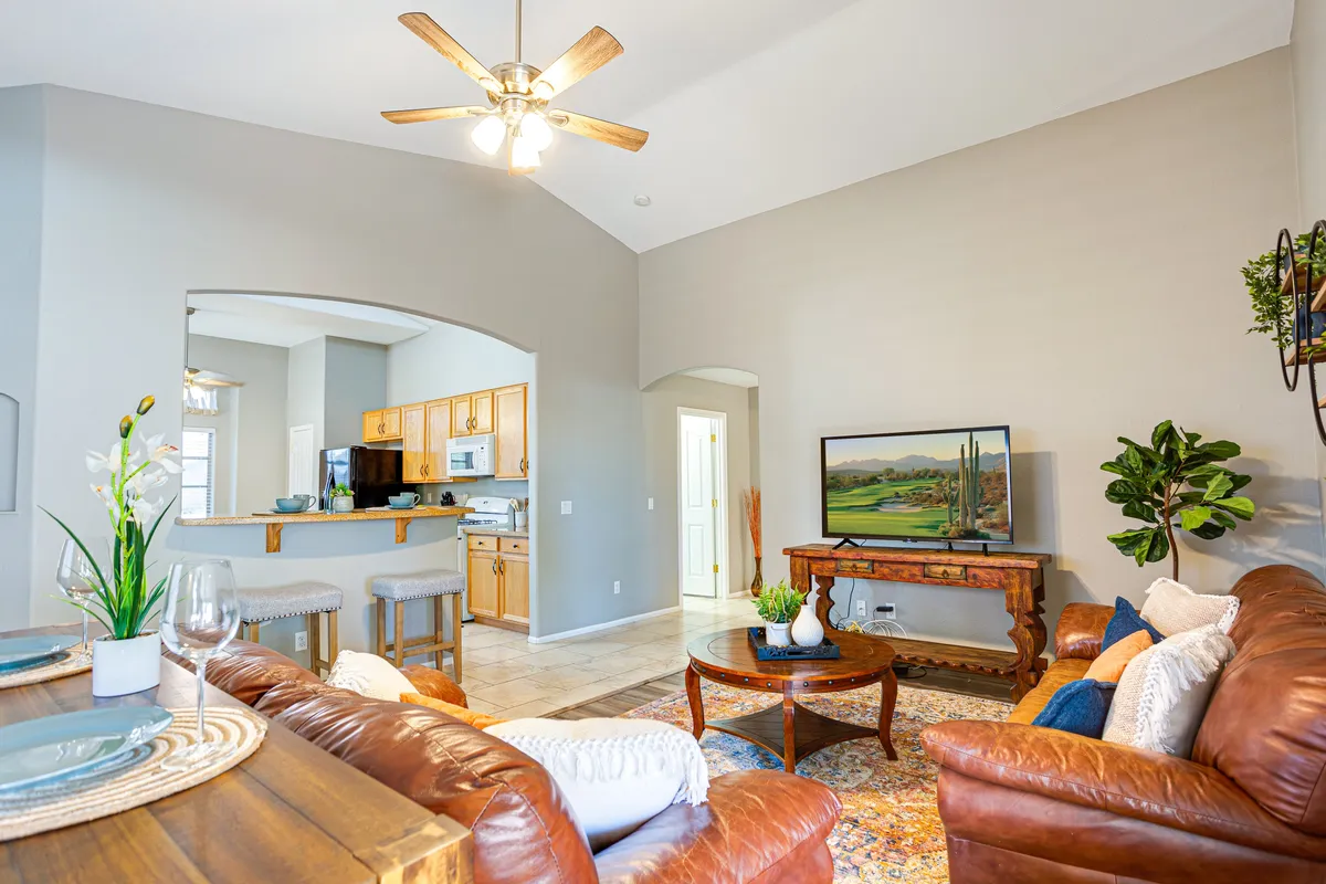 This image displays a cozy, open-concept living area that seamlessly connects a leather sofa seating zone, a dining table, a kitchen area, a flat-screen TV with landscape art, and touches of greenery with indoor plants. The room exudes warmth and is tailored for both relaxation and entertaining, with ample space for guests and family gatherings.