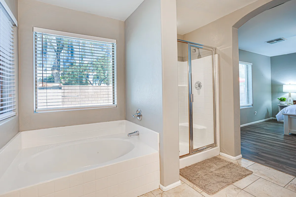 A bright, spa-like bathroom showcases a large bathtub, a separate walk-in shower, and a window letting in natural light. The spacious master bath is visible from the adjoining bedroom, highlighting double sinks and contemporary fixtures. The setting feels serene, perfect for self-care and relaxation.