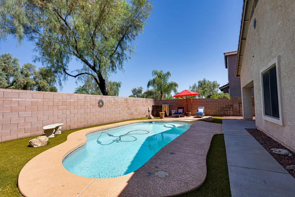 A wide exterior view of the backyard featuring the swimming pool, red umbrella, patio furniture, and lush green grass. The inviting setup includes space for outdoor dining and relaxation, with the patio visible in the background.