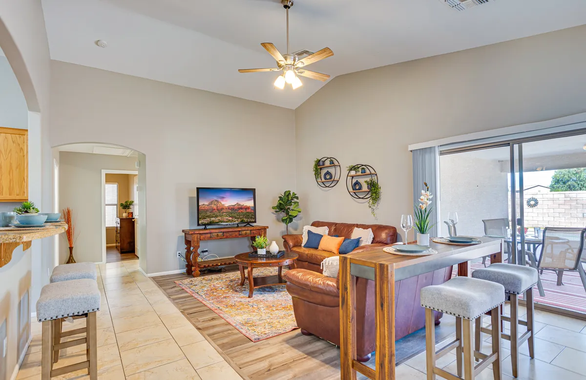 A stylish, cozy great room setting with a plush sofa, a coffee table, a large television, and a glimpse of the dining area in the background. The arrangement is conducive for relaxing, watching TV, hosting guests, and enjoying intimate dinners or game nights.